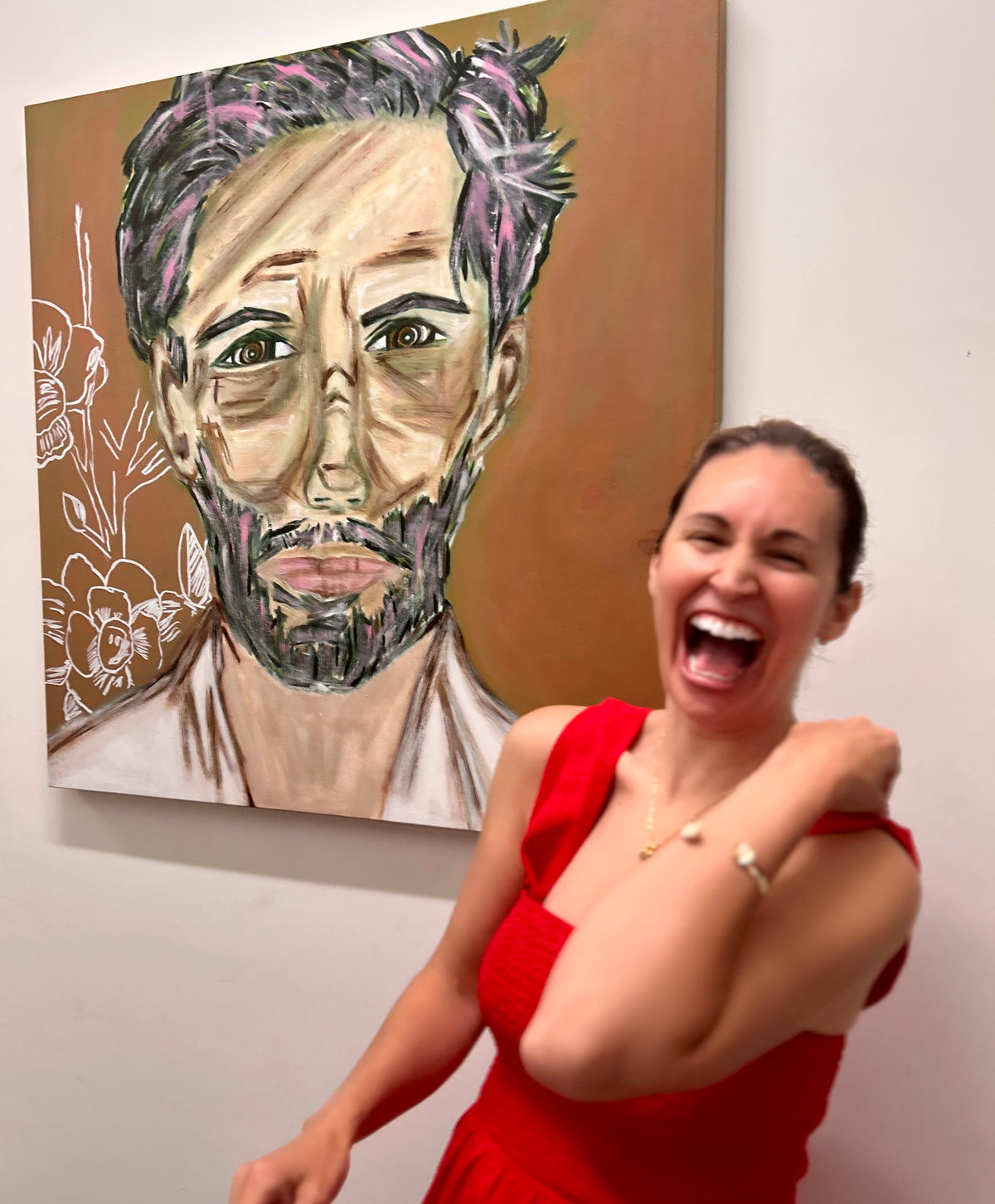 The contemporary figurative artist, Cristina Henley, in a red dress standing next to painting portraits in acrylic of a man's face on a wall. She represents meaning full arts and original narrative artwork about resilience and personal transformation.
